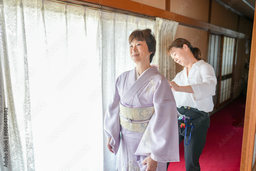 Obraz premium A woman in a kimono is getting her hair done by another woman. The scene is set in a room with a window and a curtain. Scene is calm and peaceful, as the two women are focused on their task