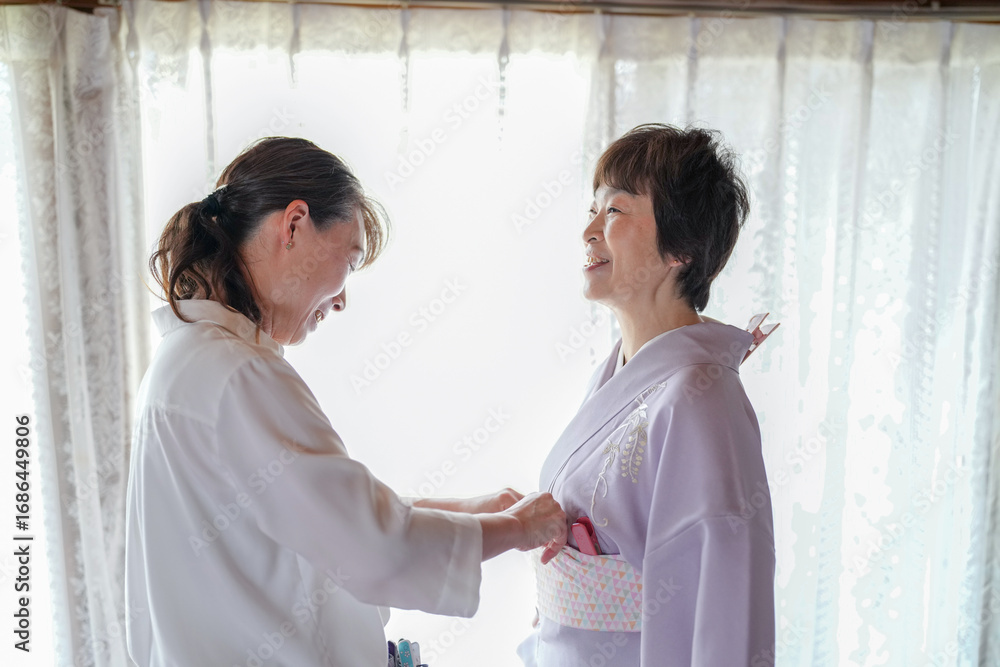 Obraz premium A woman in a kimono is being helped by a woman in a white shirt. The woman in the white shirt is helping the woman in the kimono adjust her clothing. Scene is one of care and assistance