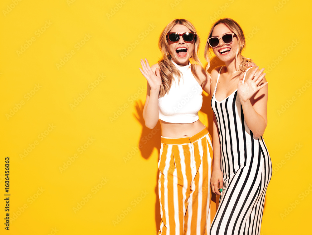 Obraz premium Two young beautiful smiling blond hipster female in trendy summer clothes. Sexy carefree women posing near yellow wall in studio. Positive models having fun. Cheerful and happy. In sunglasses