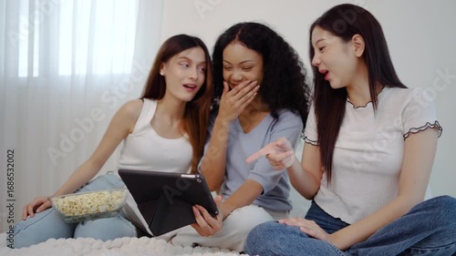 Three young asian women friends share a joyful moment together in bedroom, eagerly looking at their tablet computer and reacting with surprise and delight to the wonderful news they have just found