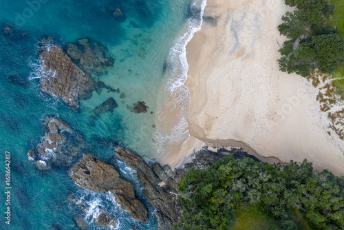 Aerial view of where the turquoise ocean meets the sandy shore near the rocks and vibrant green trees, Langs Beach, Northland Region, New Zealand.