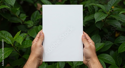 A white book is held by two hands against a green leafy backdrop