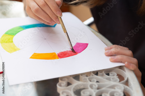Close-up of a brush applying red watercolor to a segment of a color wheel. White paper on a decorative outdoor table. Close-up view with copy space. Concept of artistic education and concentration