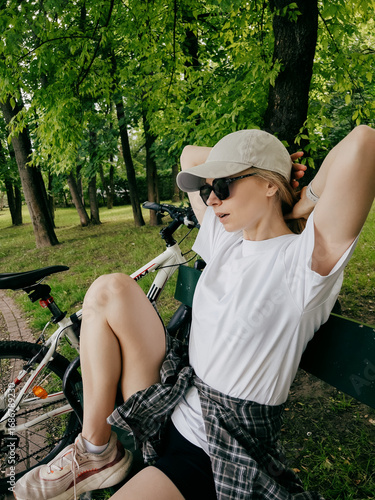 Young blonde woman cyclist wearing white t shirt, shorts, sneakers and cap is sitting on bench in green park after cycling, holding her hair with her hands and resting near her bicycle. Woman pausing