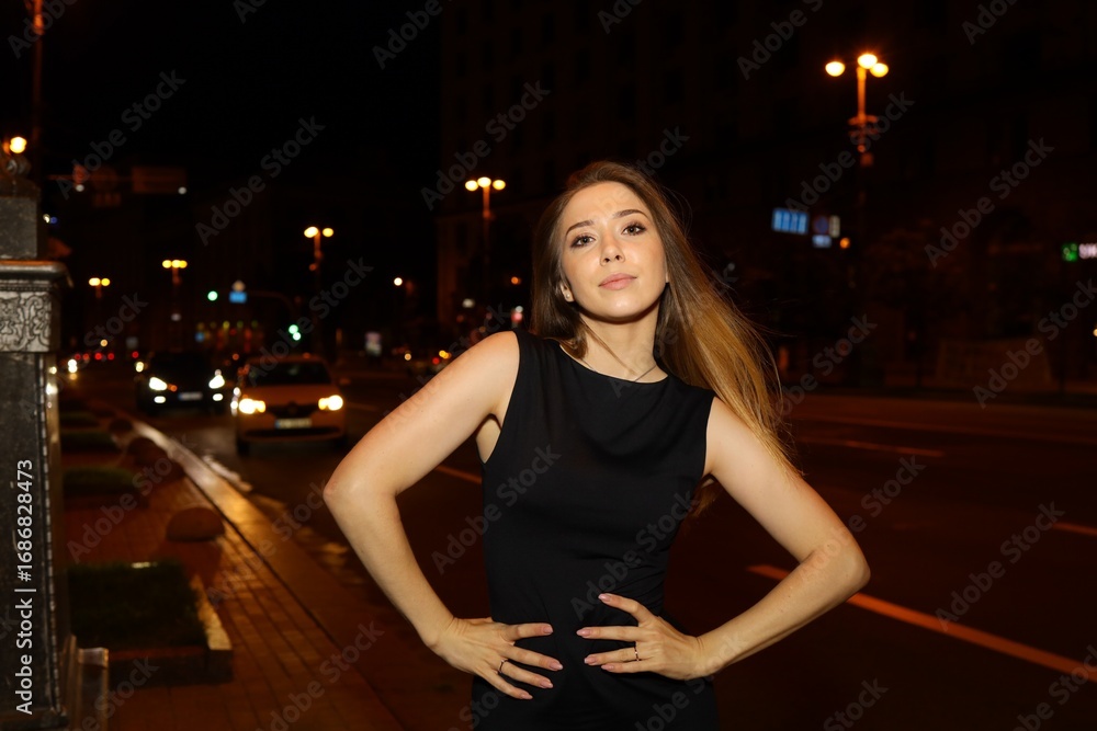 Fototapeta premium A woman is standing casually on a bustling city street at night, wearing a simple black dress.