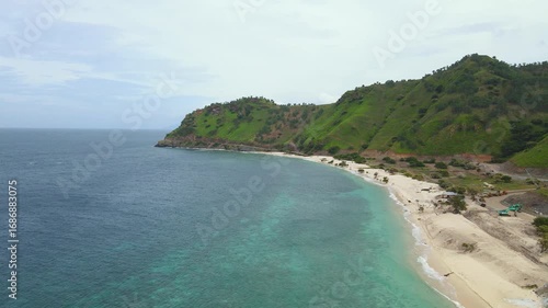 A stunning aerial view of Cristo Rei Beach in Dili, Timor-Leste. The curving white sand coastline meets clear turquoise seawater at the foot of the iconic green hills. 