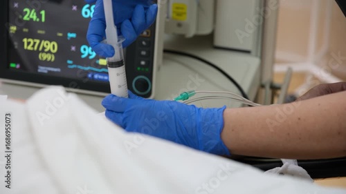 Medical professional wearing blue gloves injecting medicine with a syringe to a patient lying in a hospital bed, with vital signs monitor in the background