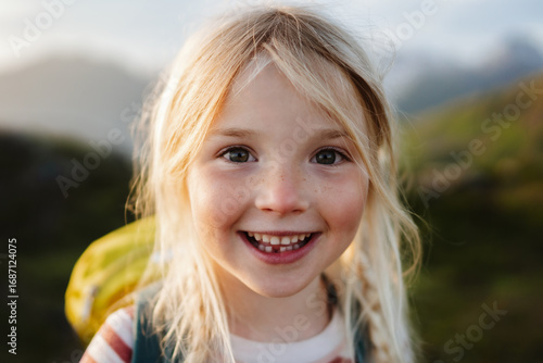 Child girl happy smiling face 5 years old kid hiking with backpack outdoor portrait looking at camera close up blonde hair family travel vacations candid emotions