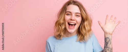 The young woman waving and laughing in a pastel pink studio portrait with tattooed arm
