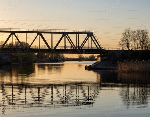 Silhouettes of cyclists crossing a steel truss bridge over a calm river during a golden spring sunset with water reflections.