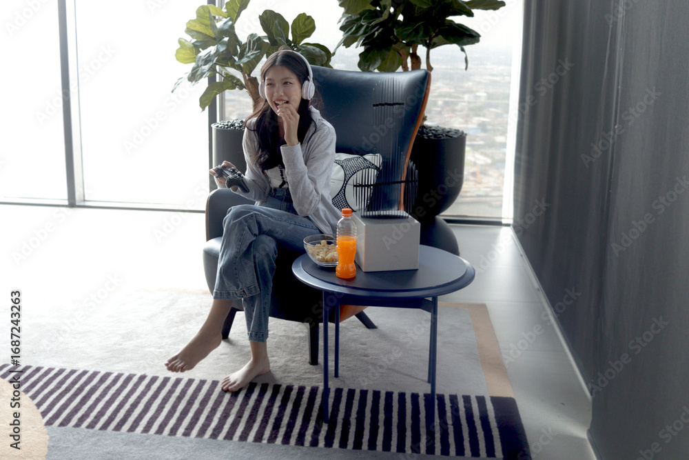 Fototapeta premium Excited Asian teenager woman with headphones holds video games joystick controller to play game while sits on sofa in living room. Happy beautiful teen girl eats snack while plays game online at home.
