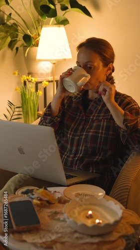 the girl is working at the computer, drinking tea. The room is dimmed, cozy and warm.