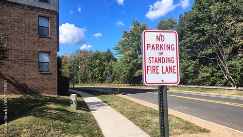 A road sign forbidding parking and stopping in the fire lane is located to the right of the brick building in front of the trees along the road.