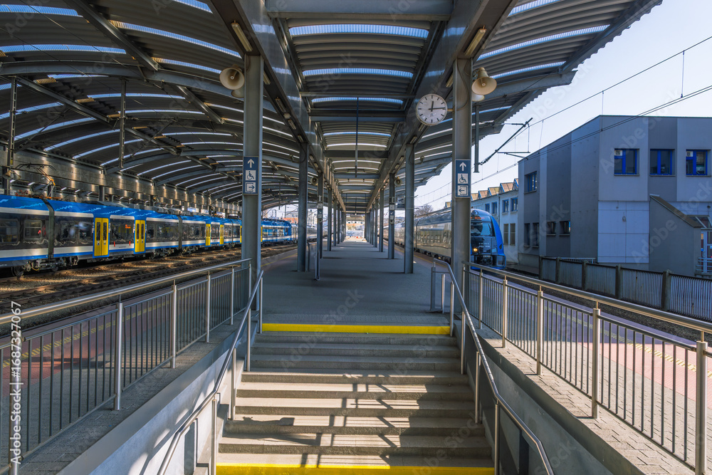 Obraz premium Train station platform with stairs leading to waiting trains under an open canopy, providing shelter and access for rail passengers.
