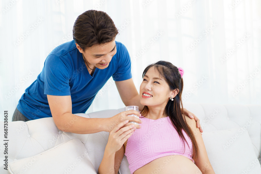 Fototapeta premium Husband caring for pregnant wife by giving her milk while she rests on sofa.
