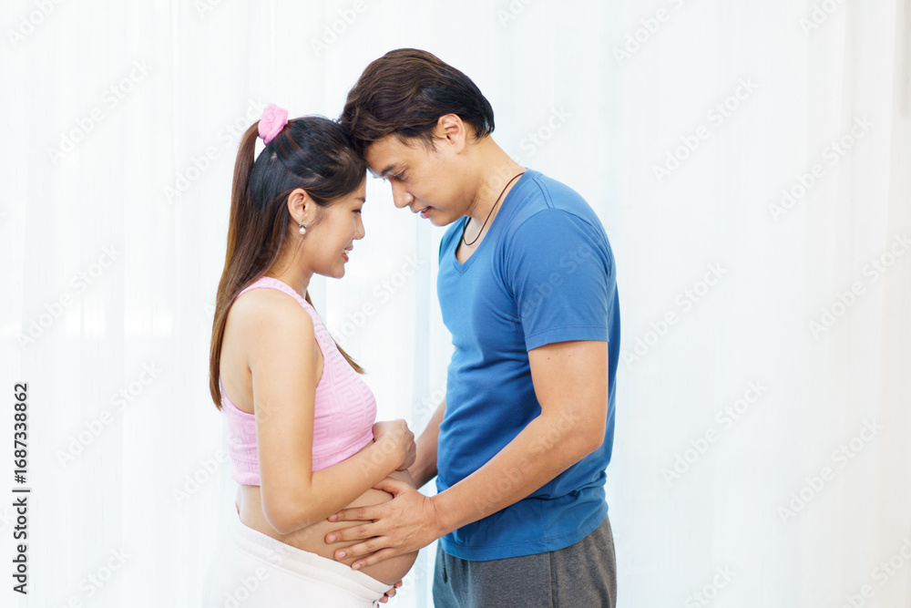 Fototapeta premium Asian husband hugging pregnant wife with hands on belly in heart shape, symbolizing love and care.
