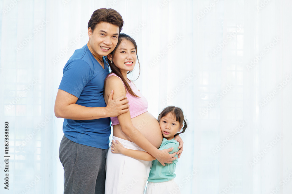 Fototapeta premium Asian family with pregnant mother, father, and daughter hugging belly with love and joy.