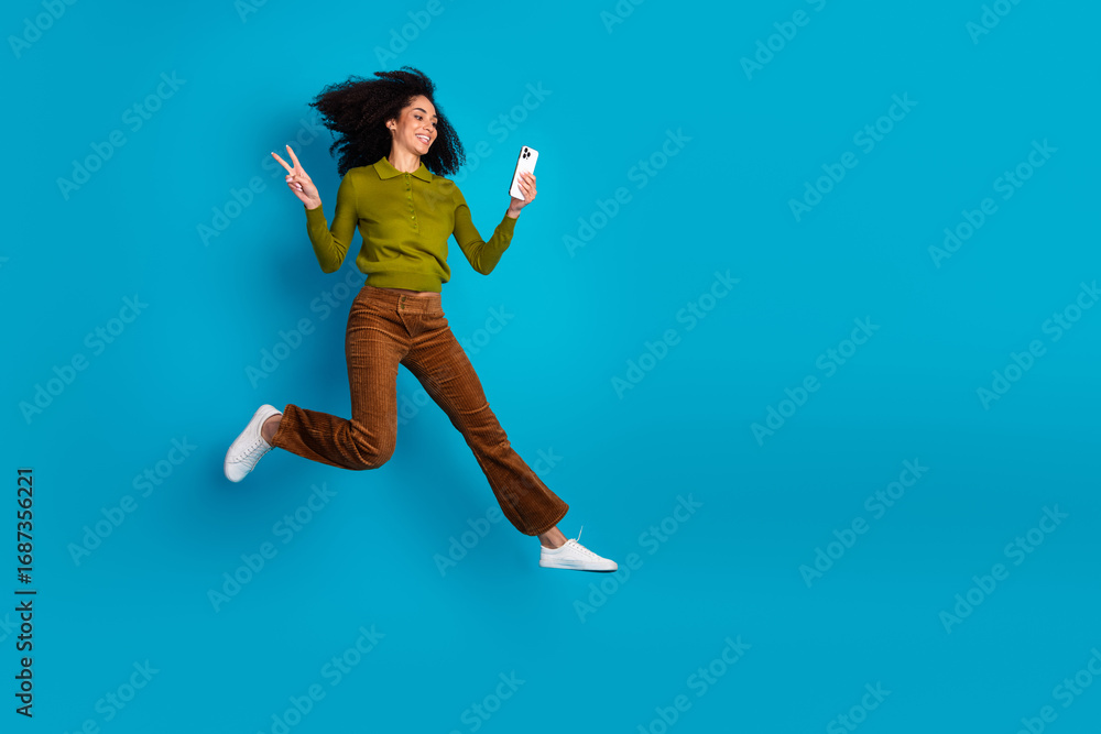 Obraz premium Energetic woman making peace sign while holding a smartphone against a blue background in casual stylish attire