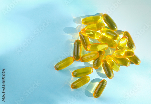 Group of yellow soft gel capsules scattered on a blue surface with natural light and shadows, symbolizing vitamins, omega-3, and dietary supplements.
