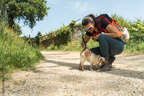 A woman with a backpack pets a cat on the Camino de Santiago route in Spain. The cat rubs against her legs and she bends down to pet it.