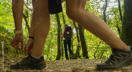 Two women are walking along the Camino de Santiago, and one of them stops to tie her shoelace. The other woman is seen carrying a large backpack, Camino de Santiago, Spain.