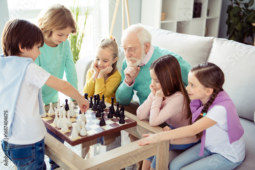 Photo of thoughtful minded grandfather in glasses playing chess with kids thinking about next move strategy at home house