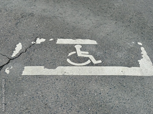 The faded accessible parking symbol is located on a cracked concrete surface within public area. An old and faded wheelchair icon is indicating an accessible parking space on cracked pavement surface