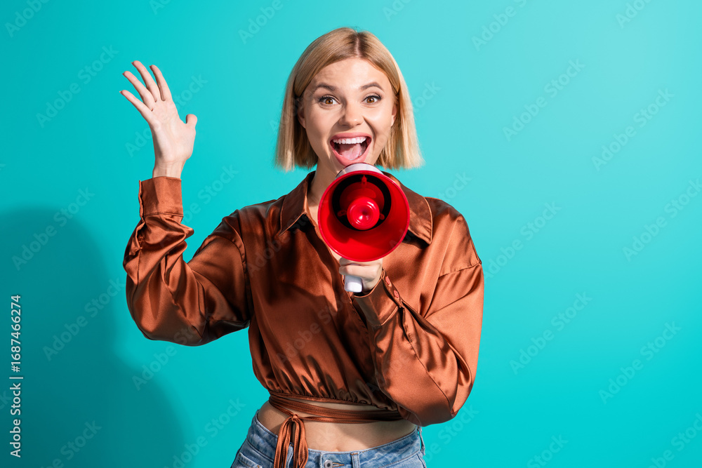 Obraz premium Youthful woman with blonde bob holding a red megaphone on turquoise background, radiating happiness and enthusiasm
