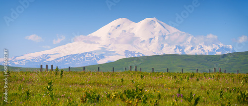 A panoramic view of the snow-capped peaks of Mount Elbrus with the green field of the Bolshoy Bermamyt plateau in the foreground, Stavropol Krai, Russia