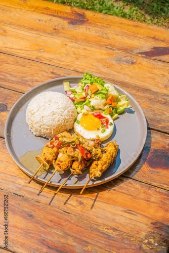 Delicious grilled chicken skewers with rice, salad, and a fried egg on a rustic wooden table