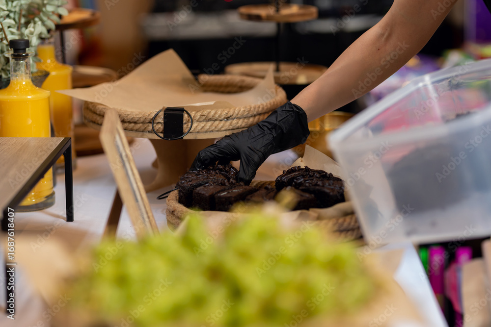 Fototapeta premium Professional caterer wearing black gloves carefully arranging chocolate brownies on serving tray, preparing gourmet dessert selection for catered event