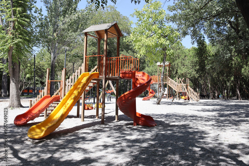 Vertical view of a wooden playground set with bright orange and yellow slides under trees. Useful for childhood, outdoor activity, park equipment, family recreation and community theme