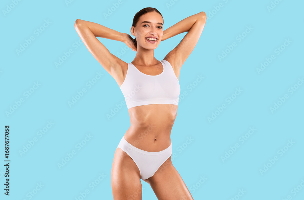 Naklejka premium Portrait of happy smiling fit woman wearing lingerie posing isolated om blue studio background, raising arms up. Female with flat belly in white underwear top bra and panties showing smooth armpits