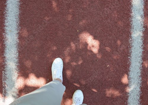 Overhead view of white sneakers standing on a red running track between white lane lines. Great for fitness motivation, jogging, sport background, active lifestyle, training, and start or finish ideas