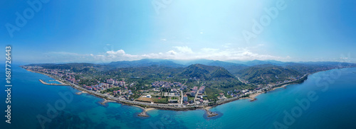 Aerial panoramic view of Arsin coastline in Trabzon, Turkey. Scenic Black Sea landscape featuring coastal settlements, deep blue water, fish farms, and green mountains in the background.