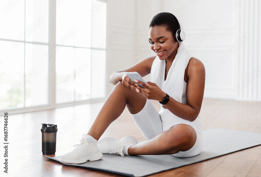 Obraz premium Happy young African woman with towel on shoulders and wireless headset sitting on fitness mat in living room, listening to music while resting, free copy space. Sports music mobile application concept