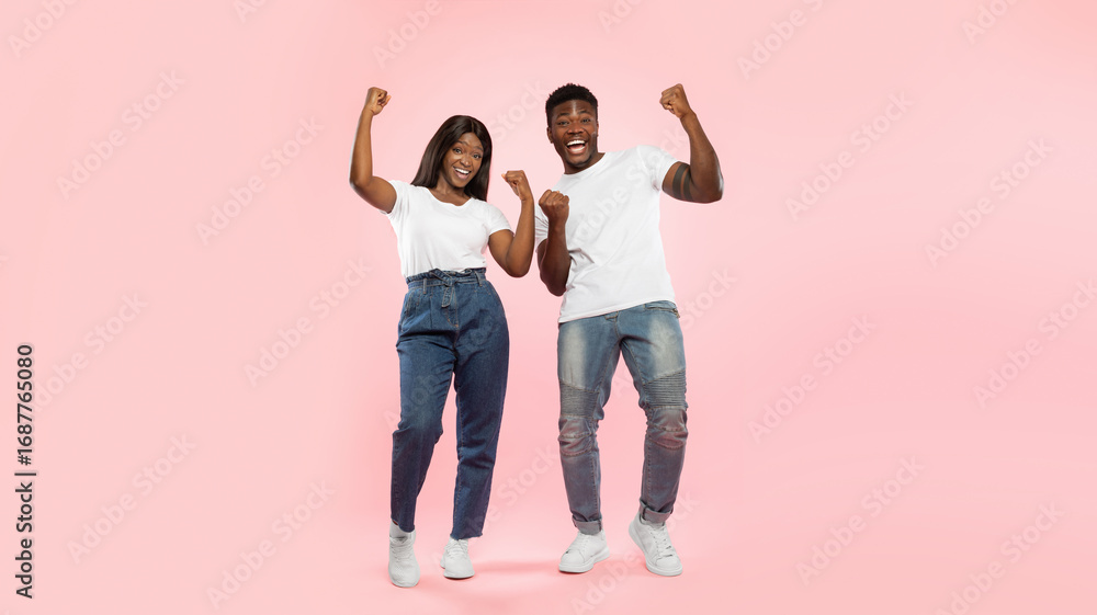 Fototapeta premium Wow, Yes. Overjoyed young black man and woman cheering and shaking clenched fists, looking at camera. Happy couple celebrating win posing standing isolated on pink studio wall, full body length