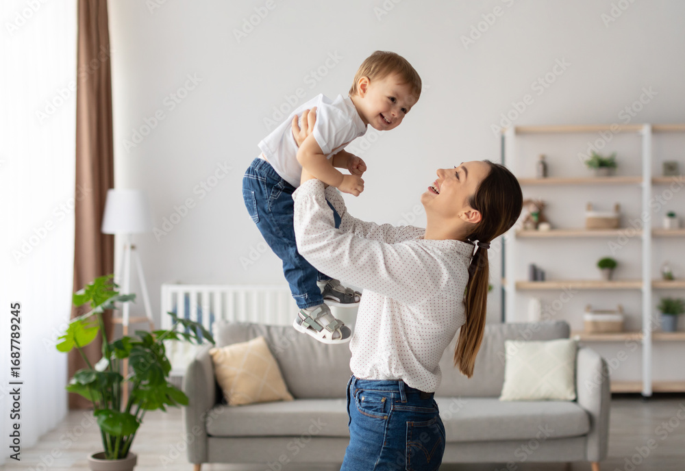 Fototapeta premium Laughing young mother playing with cute toddler son, holding funny kid pretending flying, loving happy mum lifting child boy, having fun together at home