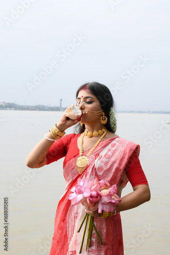 Beautiful young Indian woman, dressed in a traditional saree, with gold jewellery, playing a shankh in front of the river Ganges