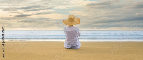 Woman in straw hat sitting on the beach at dawn, facing the sea in soft golden light.
