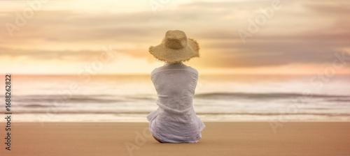 Woman in straw hat sitting on the beach at dawn, facing the sea in soft golden light.