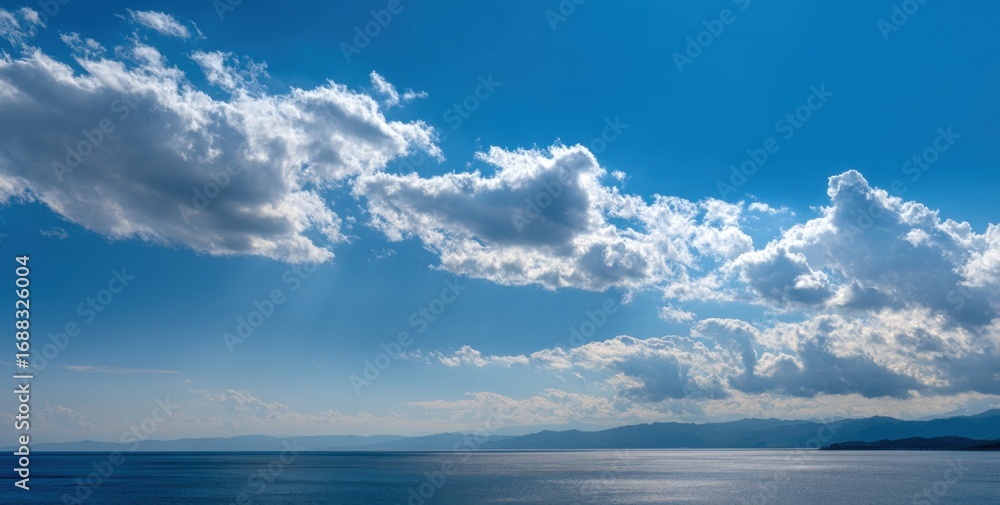 Obraz premium Panoramic view of a serene lake under a vibrant blue sky dotted with fluffy clouds. Horizon line displays distant, indistinct mountains