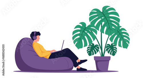 Young man wearing headphones works on a laptop while relaxing in a bean bag chair next to a large potted plant