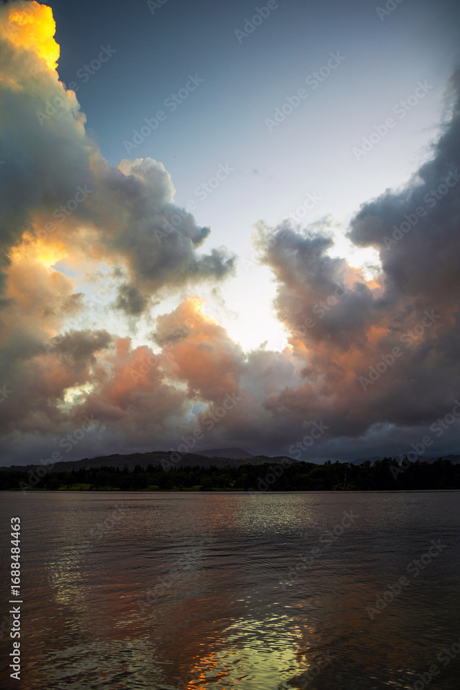 Fototapeta premium Billowing clouds above a lake, highlighted with the colours of a setting sun. Billowing clouds with warm colours of a setting sun above Lake Windermere