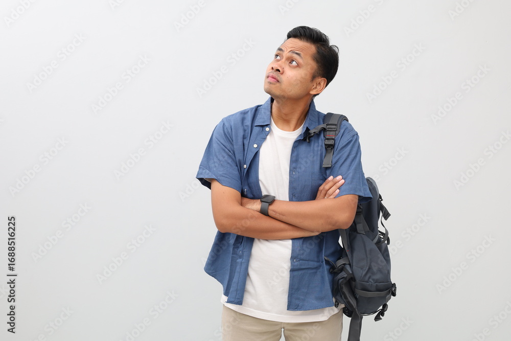 Fototapeta premium Young asian student man wearing casual clothes and backpack, standing with folded arms and looking up in empty space, isolated on white background.