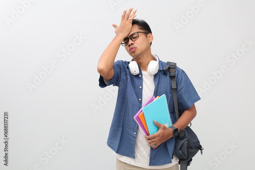 Young Asian male college student wearing casual clothes, backpack, glasses, headphones around neck while carrying books, forgetting something or making a mistake while holding forehead.