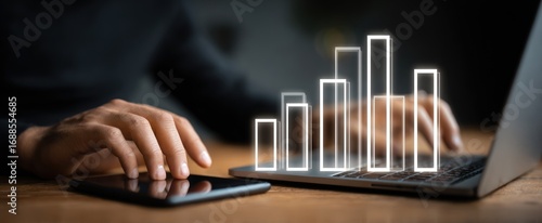 Person using a laptop and phone, with an overlaid graphic showing rising bars. Close up, hands on devices, dark clothing and background