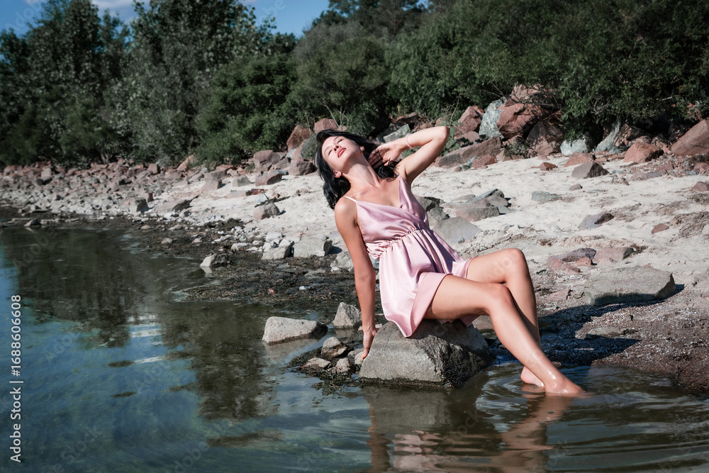 Fototapeta premium Young sexy beautiful woman is sitting on the rocks in the sea near the shore and enjoys relaxation, the warm sun and nature. Wellness, tourism, holidays, vacation and weekends.