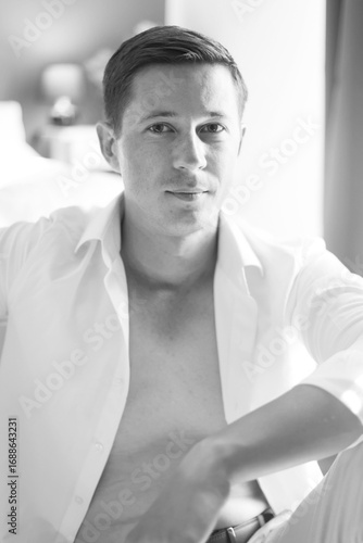 An attractive man in a white shirt is sitting near the bed. Portrait of a handsome man in a white shirt. 