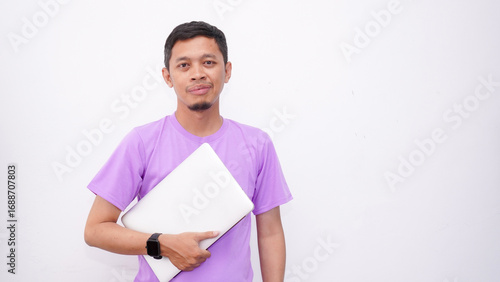 Happy asian man wearing purple t-shirt and holding laptop computer working online and looking at camera isolated on white background, online education